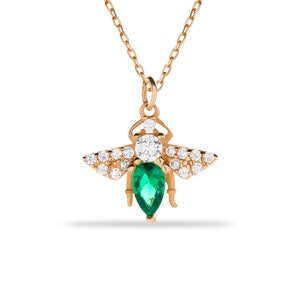 Puede incluir: Un collar de oro con un colgante de abeja con una piedra preciosa de esmeralda verde y piedras de circonita c&uacute;bica transparentes.