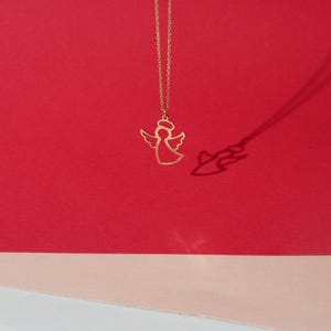May include: A gold angel pendant necklace on a red and pink background. The angel is a simple outline with wings and a halo.