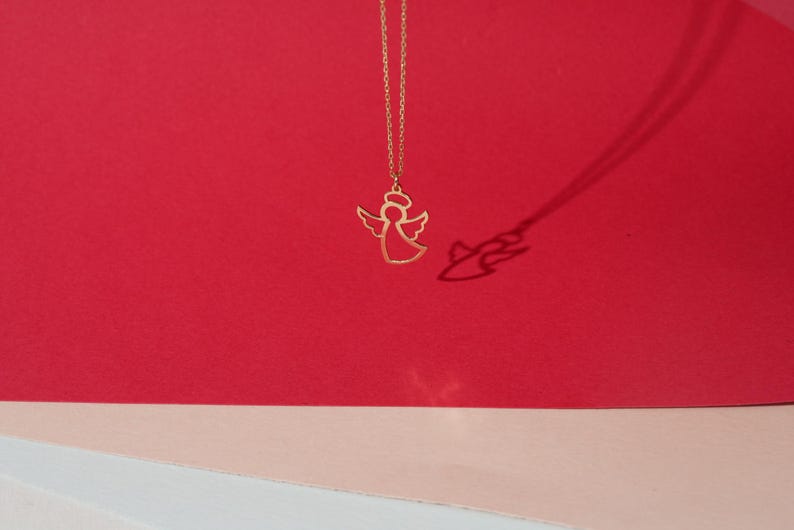 May include: A gold angel pendant necklace on a red and pink background. The angel is a simple outline with wings and a halo.