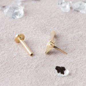 Op de afbeelding: Gouden lichaamsieraden met een schroefdraad aan het uiteinde en een platte, ronde bovenkant. De sieraden zijn gedemonteerd en tonen de twee delen afzonderlijk.