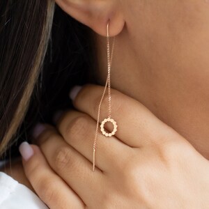 May include: A delicate gold-colored earring with a chain and a small, circular pendant. The pendant is adorned with tiny, evenly spaced beads. The earring is shown dangling from an ear, showcasing its elegant design.