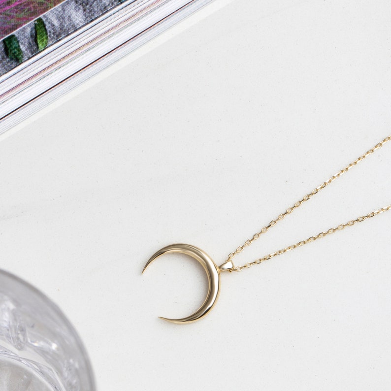 Puede incluir: Un collar con un colgante de media luna de oro en una cadena delicada. El colgante es un dise&ntilde;o simple y elegante.