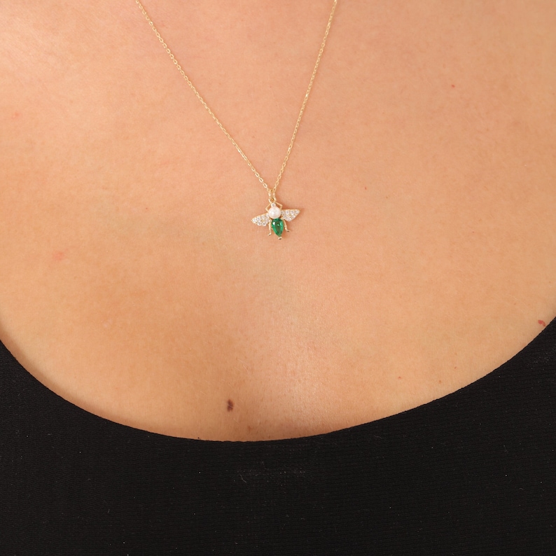 Puede incluir: Un collar de cadena dorada con un peque&ntilde;o colgante de abeja. La abeja tiene un cuerpo de piedra preciosa verde y detalles de cristal blanco.