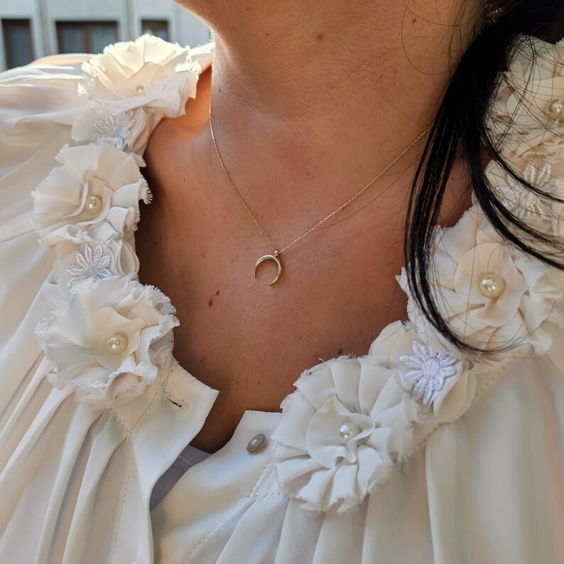 Puede incluir: Una mujer con una blusa blanca con delicadas flores de tela blanca y un collar de media luna de oro.