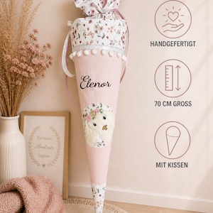 Könnte beinhalten: Eine rosa Schultüte mit Blumenmuster und dem aufgedruckten Namen "Elenor". Die Tüte ist mit weißen Pompons und einem Blumendesign am oberen Rand verziert. Der Text "70 CM GROSS" ist sichtbar.