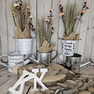 Könnte beinhalten: Drei verzinkte Gießkannen aus Metall mit verschiedenen personalisierten Botschaften. Die Kannen sind mit Trockenblumen gefüllt und auf einer Holzscheibe platziert. Die Botschaften auf den Kannen lauten "Lieb Ralf alle Liebe zu 60. Geburtstag", "Friends are the family we choose" und "Mama Jedine Sascha IVIA".
