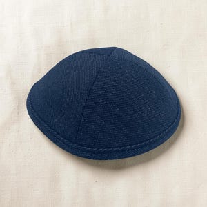 Può includere: Una kippah in denim blu scuro caratterizzata da una classica struttura a quattro pannelli con cuciture visibili lungo le giunture e un bordo circolare rinforzato. Il tessuto presenta una distinta trama intrecciata tipica del denim.