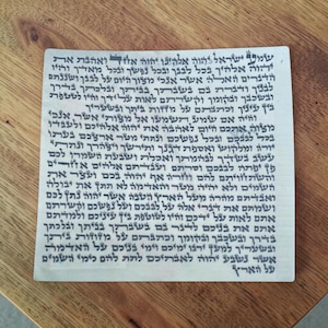 May include: A square parchment featuring dense, dark Hebrew calligraphy printed in black ink. The text consists of multiple lines of traditional religious script, covering the entire surface of the off-white, textured material from edge to edge.