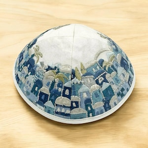 Può includere: Kippah ricamata con un design dettagliato di paesaggio urbano nei toni del blu, grigio e oro. La kippah ha una base bianca e un bordo bianco. Il design raffigura edifici, palme e altri elementi architettonici.