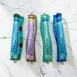 May include: Four vertical mezuzah cases featuring clear glass tubes with blue Hebrew text scrolls inside. Each case has a colored glass backing in green, brown, light green, and blue, with a matching colored glass sphere at the top of each tube.