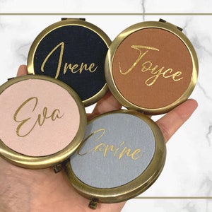 Puede incluir: Cuatro espejos compactos con ribete dorado y cubiertas de tela de diferentes colores. Los espejos están personalizados con los nombres "Irene", "Joyce", "Eva" y "Carine" en letras doradas.