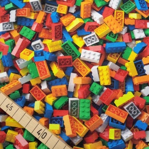 Könnte beinhalten: Ein farbenfrohes Stoffmuster mit einer dichten Anordnung von Spielzeug-Bausteinen in verschiedenen Farben, darunter Rot, Grün, Gelb, Blau, Orange und Weiß. Die Bausteine sind unterschiedlich groß und geformt und erzeugen ein verspieltes Design. Ein Holzlineal mit Markierungen ist sichtbar.