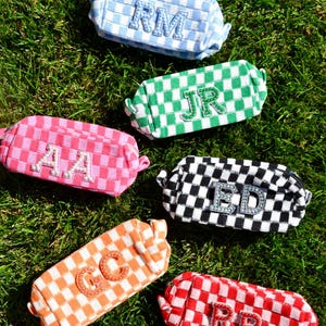 Peut inclure: Plusieurs trousses de maquillage rectangulaires à carreaux, dans des couleurs variées : bleu, vert, rose, noir, orange et rouge. Chaque trousse arbore des initiales brodées, comme "RM", "JR" et "ED", dans une couleur contrastante.
