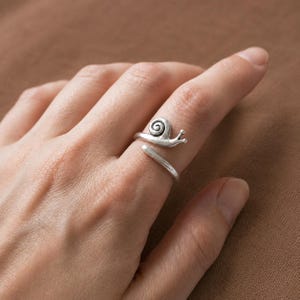 May include: A silver-toned open ring featuring a sculpted snail design. The snail is positioned at one end of the band, with its shell showing a spiral pattern and two small antennae extending forward. The band is smooth and wraps around the finger.