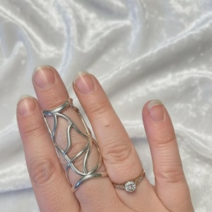 May include: A silver finger armor ring and a gold ring with a clear stone are displayed. The finger armor ring has an open, web-like design. The gold ring features a small, round, clear stone.