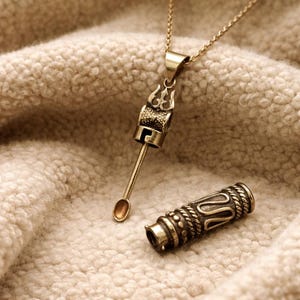 May include: A gold-coloured pendant necklace with an intricate, ornate design. The pendant has a cylindrical body with decorative patterns and a small spoon-like attachment. The necklace is displayed on a textured, beige surface.