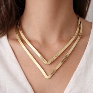 Könnte beinhalten: Eine goldfarbene Halskette mit doppeltem V-förmigem Design. Die Halskette wird um den Hals getragen und hat eine gehämmerten Textur. Der Schmuck ist aus Metall und ein Statement-Stück.
