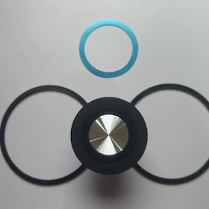 Puede incluir: Un objeto negro y plateado con un diseño circular, posiblemente un altavoz o un botón, está rodeado por tres anillos negros circulares y un anillo azul. El objeto tiene un centro plateado metálico.