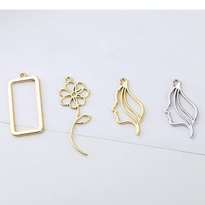 May include: Four metal charms in gold and silver tones. The charms include a rectangular frame, a flower, and two profile silhouettes. The charms are designed for jewelry making or crafting.
