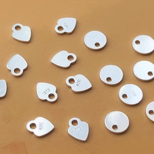 Puede incluir: Una colecci&oacute;n de 14 dijes de metal plateado, incluyendo dijes redondos y en forma de coraz&oacute;n. Algunos dijes tienen un agujero en el centro, mientras que otros tienen dos agujeros. Los dijes est&aacute;n en blanco, a excepci&oacute;n de algunos que tienen el n&uacute;mero "925" grabado.