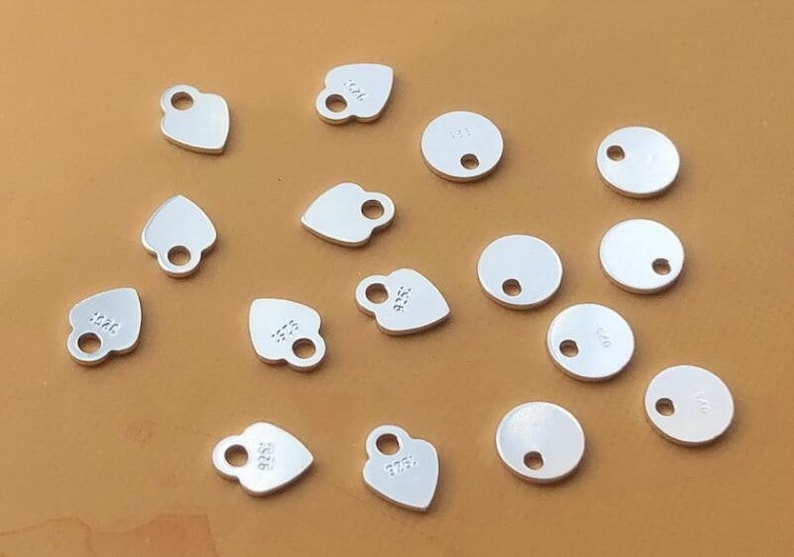 Puede incluir: Una colecci&oacute;n de 14 dijes de metal plateado, incluyendo dijes redondos y en forma de coraz&oacute;n. Algunos dijes tienen un agujero en el centro, mientras que otros tienen dos agujeros. Los dijes est&aacute;n en blanco, a excepci&oacute;n de algunos que tienen el n&uacute;mero "925" grabado.