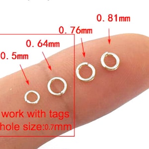 Puede incluir: Cuatro anillos de salto de oro de diferentes tama&ntilde;os, 0,5 mm, 0,64 mm, 0,76 mm y 0,81 mm, en un dedo. El texto "work with tags hole size: 0.7mm" est&aacute; debajo de los anillos.