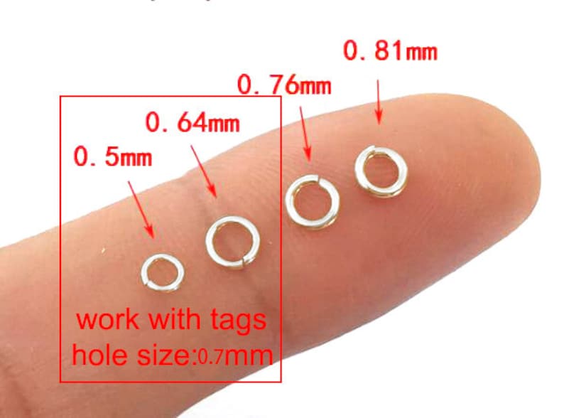 Puede incluir: Cuatro anillos de salto de oro de diferentes tama&ntilde;os, 0,5 mm, 0,64 mm, 0,76 mm y 0,81 mm, en un dedo. El texto "work with tags hole size: 0.7mm" est&aacute; debajo de los anillos.