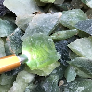 May include: A close-up of a pile of green and white jade stones. The stones are rough and have a natural, unpolished look. The stones are illuminated by a flashlight, highlighting their translucent quality.