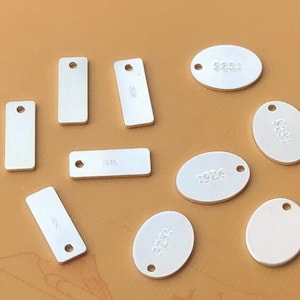 Puede incluir: Diez etiquetas de metal plateadas con un agujero para colgar. Las etiquetas son de varias formas, incluyendo rectangulares y ovaladas. Algunas etiquetas tienen el n&uacute;mero "925" grabado.