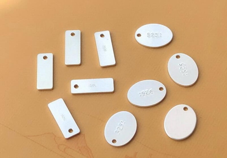 Puede incluir: Diez etiquetas de metal plateadas con un agujero para colgar. Las etiquetas son de varias formas, incluyendo rectangulares y ovaladas. Algunas etiquetas tienen el n&uacute;mero "925" grabado.