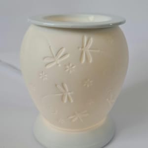 May include: Off-white ceramic essential oil diffuser with embossed dragonfly and floral designs. The diffuser has a light grey rim and base. The dragonfly design is repeated around the body of the diffuser.