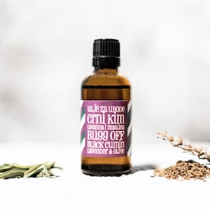 May include: A brown glass bottle with a black cap and a label that reads "uLje za uBoDe crni kim Lavanda i maslina BUgg OFF BLACK cumin Lavender & Olive". The bottle is sitting on a white surface with some dried herbs and seeds.