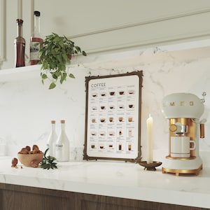 Op de afbeelding: Een witte en gouden Smeg espressomachine met een glazen kan en een witte kop op het aanrecht. Achter de machine hangt een ingelijst poster met de tekst "COFFEE THE ESSENTIALS" en een schema van verschillende koffiedrankjes. Er staan twee doorzichtige glazen flessen met witte etiketten en een kom met walnoten op het aanrecht.