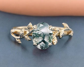 May include: A gold-colored ring featuring an oval-cut gemstone with green inclusions. The ring's band is adorned with leaf-like details on each side of the gemstone, creating a nature-inspired design. The ring is set against a dark blue background.