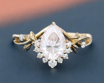May include: A gold-toned ring featuring a large, pear-shaped, clear gemstone as the centerpiece. The ring is adorned with smaller, round, clear gemstones and delicate leaf-like details on the band, creating an elegant and nature-inspired design.