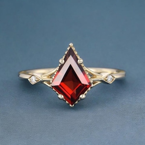May include: A gold ring with a striking, kite-shaped, deep red gemstone. The ring has a delicate design with small diamonds either side of the central stone. The band is a simple, polished gold.