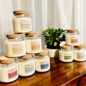 May include: A collection of glass jar candles with cork lids, each labeled with a different scent. Scents include "Lemongrass Sage," "Cake Batter," "Citrus & Sage," "Mandarin Driftwood," "Under the Mistletoe," and "Lavender Chamomile." The candles are arranged on a wooden surface.