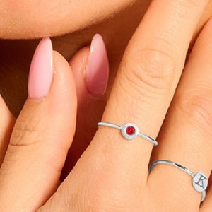 Op de afbeelding: Twee zilveren ringen aan een vinger. De ring aan de wijsvinger heeft een kleine rode edelsteen. De ring aan de middelvinger heeft een zilveren letter "K" erop.