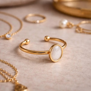 May include: Gold-toned open cuff bracelet with a white, glittery oval center stone. Other gold jewelry, including necklaces and rings, are in the background. The jewelry is displayed on a neutral surface.