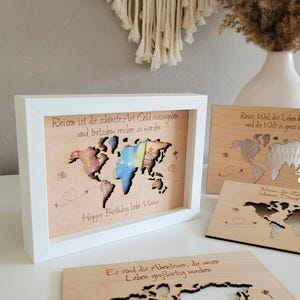 Könnte beinhalten: Ein gerahmter Weltkarte aus Holz mit weißem Rahmen. Die Karte ist lasergeschnitten mit farbigen Akzenten und deutschem Text. Der Text lautet "Reisen ist die schönste Art Geld auszugeben und trotzdem reicher zu werden" und "Happy Birthday liebe Mami".