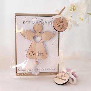 Könnte beinhalten: Ein Holzengel-Ornament mit einem Herz-Ausschnitt, dem Namen "Charlotte" und dem Text "Der immer... du ihn brauchst" auf einer Karte. Das Ornament ist mit einem Holzanhänger mit der Aufschrift "zur Taufe" verpackt.