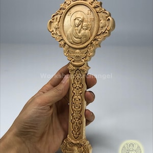 May include: A carved wooden Orthodox cross featuring a central oval relief of the Virgin Mary and child. The cross is intricately detailed with ornate carvings, including angels and floral patterns. The cross is held in a hand.
