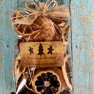 May include: A clear cellophane bag tied with raffia and lace ribbon, filled with dried orange slices, spices, and a label that reads "Winters Garden Boutique". The label features a star, tree, and gingerbread man design.