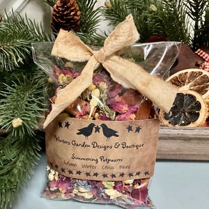 May include: A clear plastic bag filled with dried flowers, cinnamon sticks, and other potpourri ingredients. The bag is tied with a brown ribbon and has a brown paper label that reads "Winters Garden Designs & Boutique, Simmering Potpourri, Autumn Winter Citrus Blend".