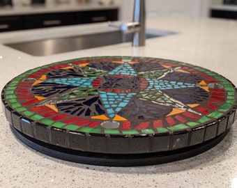 May include: A round mosaic tile trivet with a black base. The mosaic design features a starburst pattern in shades of blue, green, red, orange, and yellow.