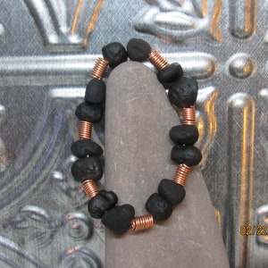May include: A handmade bracelet featuring black, irregularly shaped beads strung together with copper wire. The bracelet is displayed on a gray stone against a textured metal background. The beads appear to be natural stones.