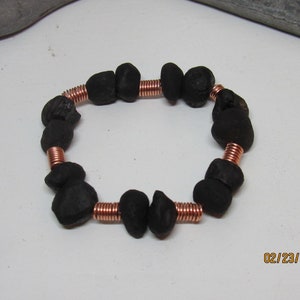 May include: A handmade bracelet featuring dark, irregularly shaped black beads strung on copper wire. The beads are spaced apart by coiled copper wire segments, creating a unique, textured look. The bracelet is displayed on a white surface.