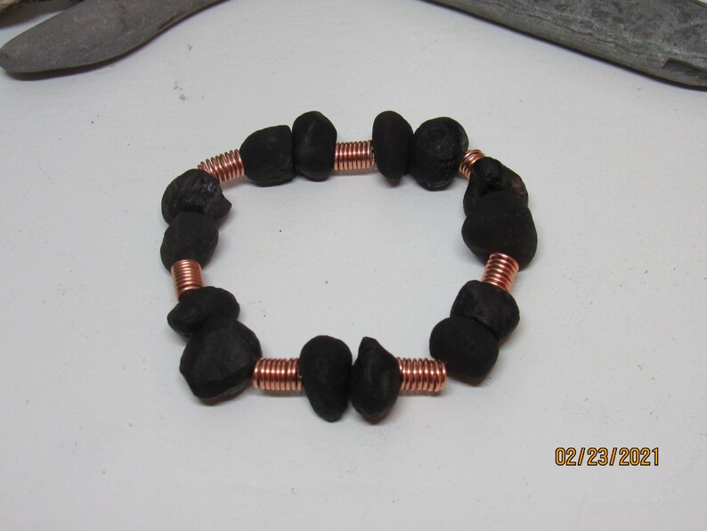May include: A handmade bracelet featuring dark, irregularly shaped black beads strung on copper wire. The beads are spaced apart by coiled copper wire segments, creating a unique, textured look. The bracelet is displayed on a white surface.
