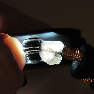 May include: Close-up of two small, irregularly shaped stones, one black and white striped, the other translucent. A copper coil and black material are attached. The date "02/23/2021" is visible.