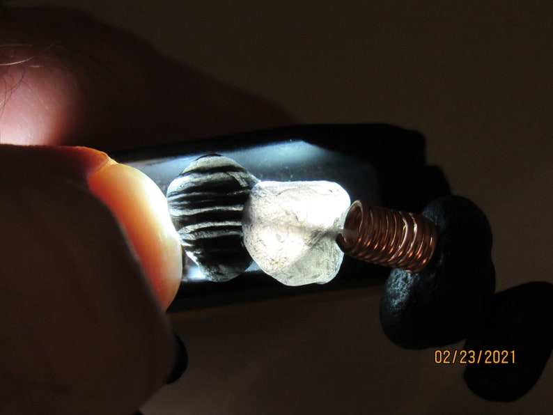 May include: Close-up of two small, irregularly shaped stones, one black and white striped, the other translucent. A copper coil and black material are attached. The date "02/23/2021" is visible.